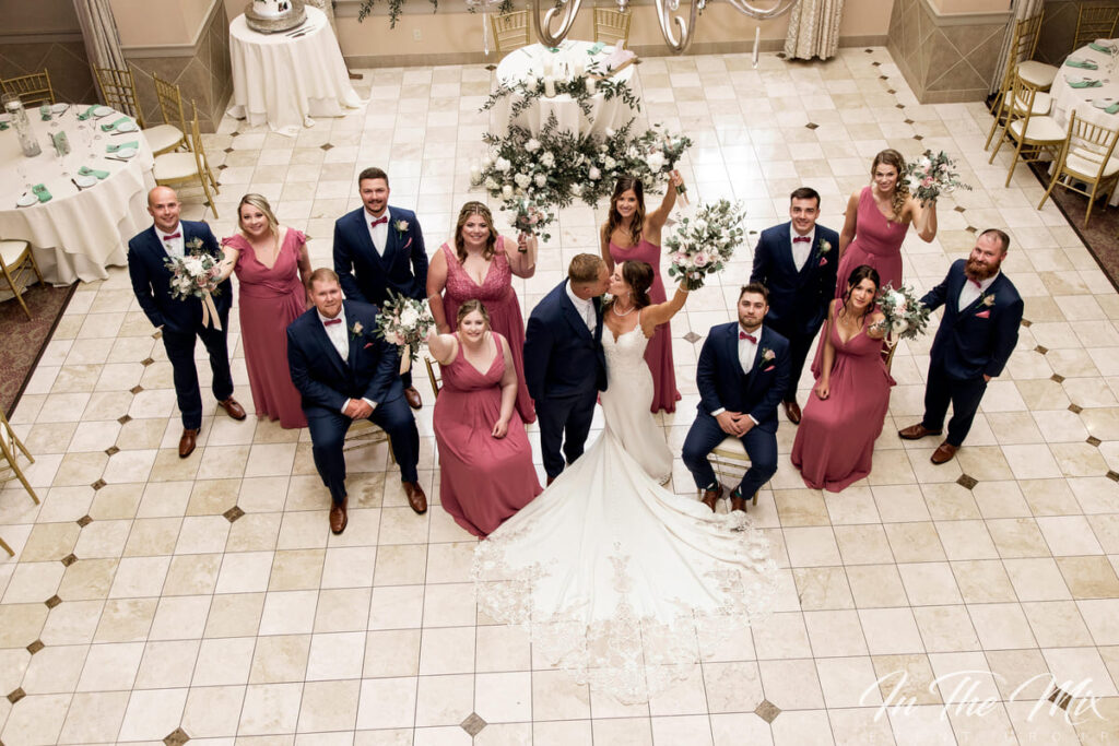 Wedding party celebration in elegant ballroom, bride and groom at center, surrounded by bridesmaids in burgundy dresses and groomsmen in navy suits, festive atmosphere with floral arrangements and banquet tables in background.