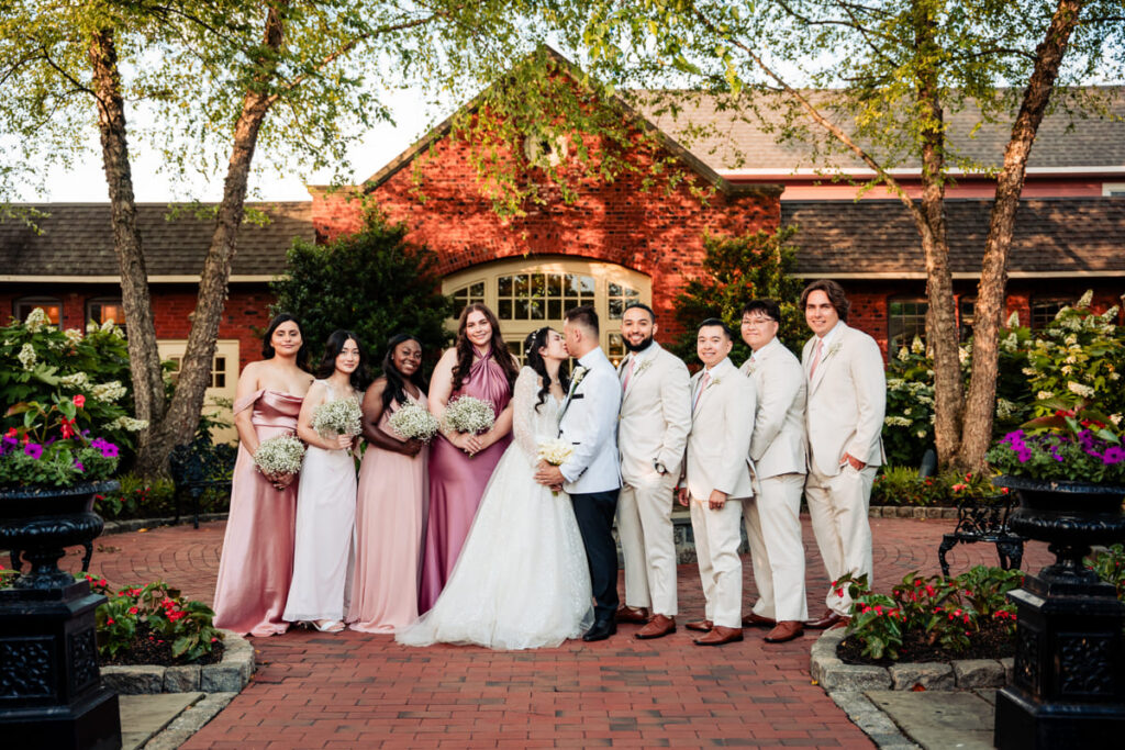 Wedding party posing outdoors at Talamore Weddings in Ambler, featuring the bride and groom at the center, surrounded by bridesmaids in blush and white dresses, and groomsmen in light suits, with lush gardens and a red brick venue in the background.